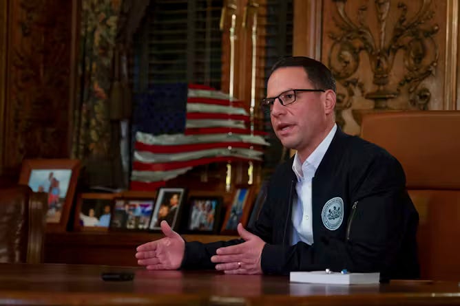 Pennsylvania Gov. Josh Shapiro’s first budget, in 2023, was not fully passed until mid-December. AMan in suit jacket and white shirt and wearing glasses sits at table with American flag in background