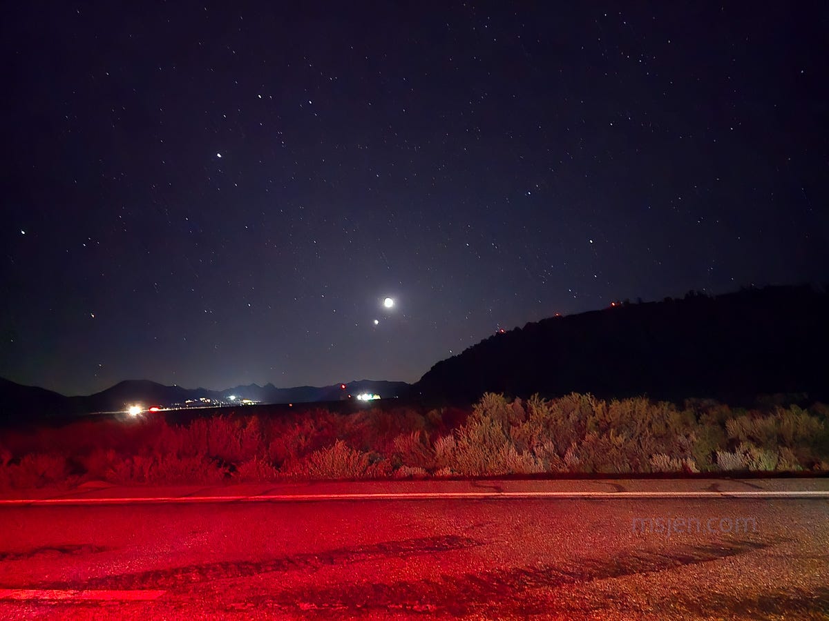 A night sky photo of the Moon and Venus conjunction with lots of road lights and red brake lights illuminating the foreground sagebrush. Photo by Jenifer Hanen, aka Ms. Jen.