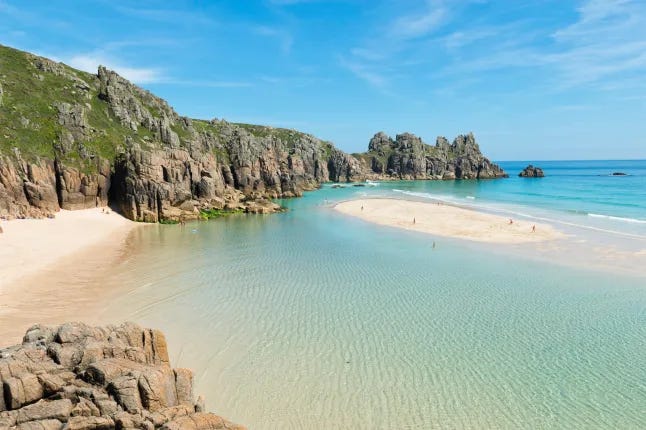 View of Pedn Vounder Beach in Cornwall at low tide, with turquoise water, golden sand, and rocky cliffs surrounding the cove. A few small, distant figures are visible on the sandbar. View of Pedn Vounder Beach in Cornwall at low tide, with turquoise water, golden sand, and rocky cliffs surrounding the cove. A few small, distant figures are visible on the sandbar.