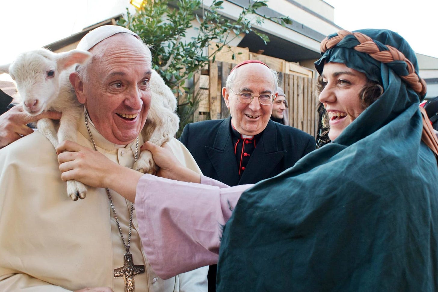 Pope Francis laughs as a woman dressed as a shepherd drapes a lamb across his shoulders at a living Nativity scene, with a smiling cardinal behind him.