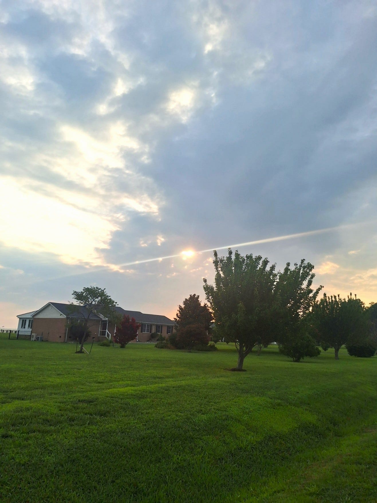 Morning sunlight breaks through gray clouds over a peaceful country lawn and trees, reflecting the quiet ways God's presence becomes visible in everyday life.