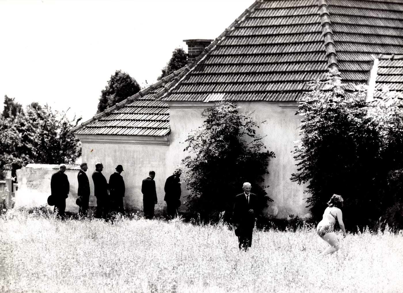 Black-clad men from a funeral procession relieve themselves against a wall, as a woman cavorts in the grass in a swimsuit.