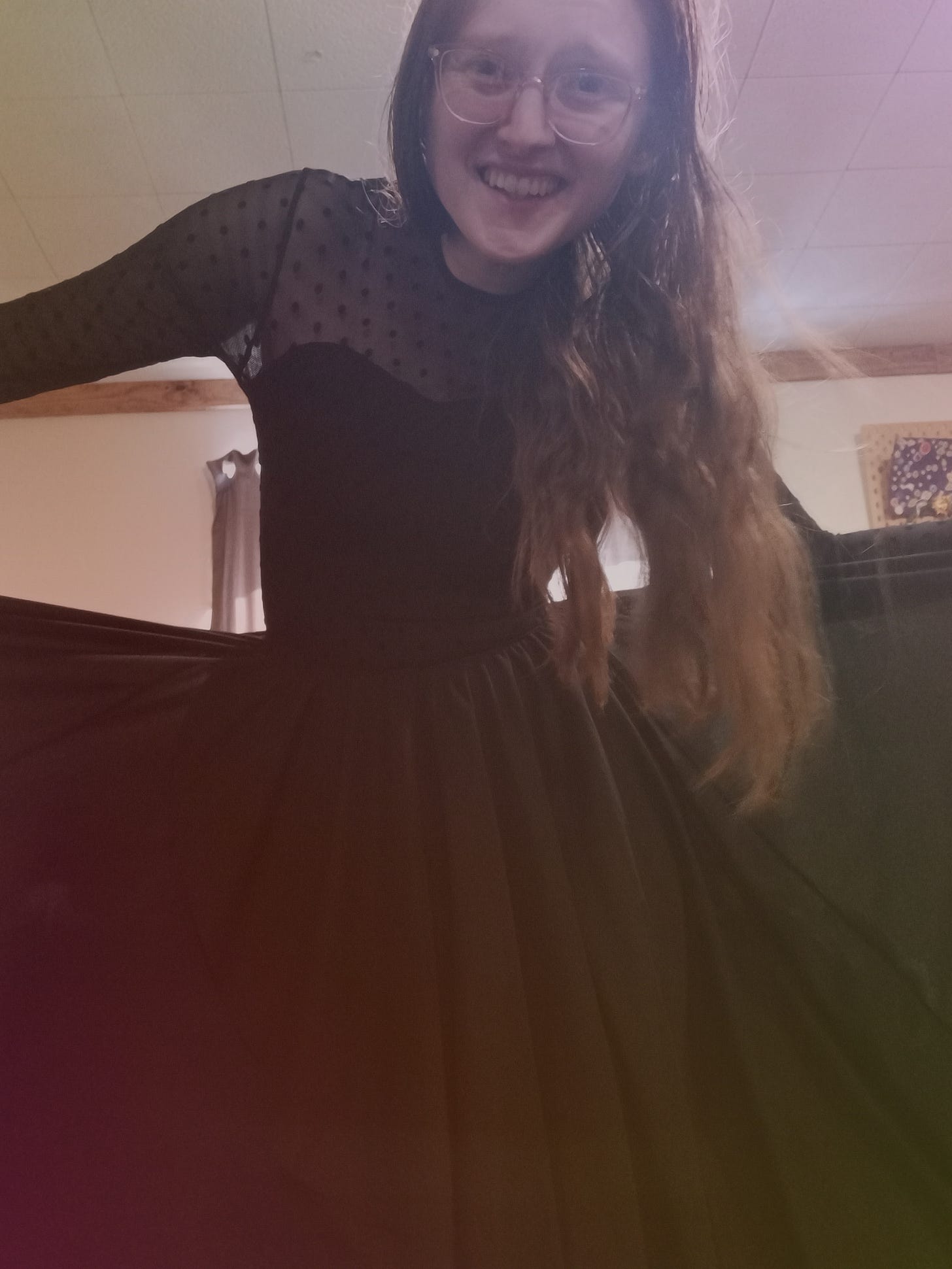 Kandi Zeller (She/Her), a white woman with long reddish brown hair, smiles at the camera while wearing a flowy black dress with a sweetheart neckline and a bodice covered in mesh with polka dots. Kandi Zeller (She/Her), a white woman with long reddish brown hair, smiles at the camera while wearing a flowy black dress with a sweetheart neckline and a bodice covered in mesh with polka dots.