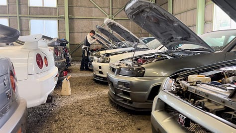 A collection of photos showing dozens of white, blue and green Nissan R34 Skyline GT-R sports cars crammed into a dimly-lit warehouse with gray walls and green support beams with a gravel floor.