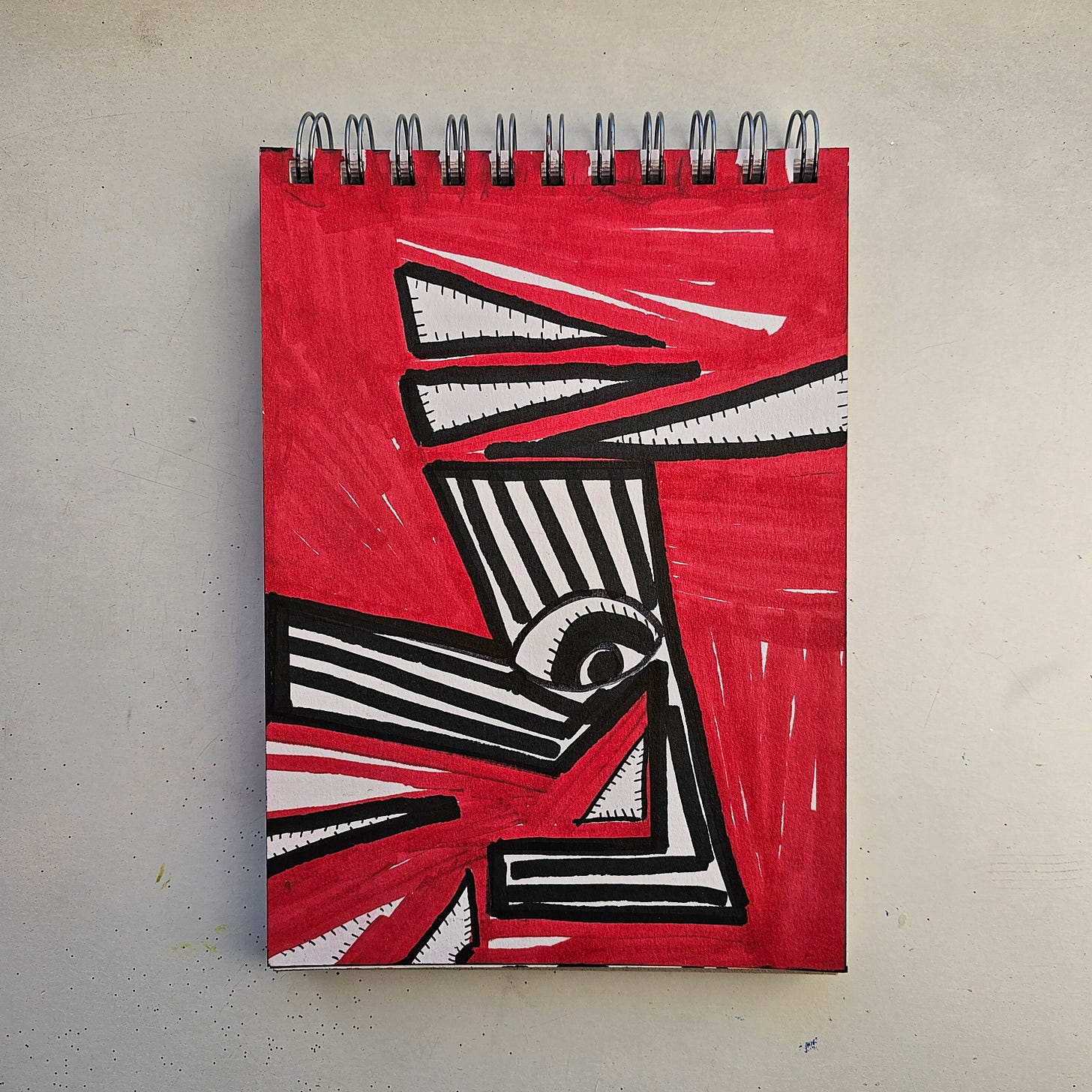 Alt Text: A sketchbook drawing by artist FatbikeHero (b. 1983) featuring a bold abstract design in red, black, and white. The composition includes sharp geometric shapes, striped patterns, and a prominent single eye, set against a saturated red marker background. The style is graphic and high-contrast, evoking kinetic energy. Alt Text: A sketchbook drawing by artist FatbikeHero (b. 1983) featuring a bold abstract design in red, black, and white. The composition includes sharp geometric shapes, striped patterns, and a prominent single eye, set against a saturated red marker background. The style is graphic and high-contrast, evoking kinetic energy.
