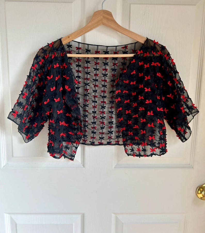 May include: A black sheer bolero with red bows embroidered all over. The bolero has short sleeves and a scoop neckline.