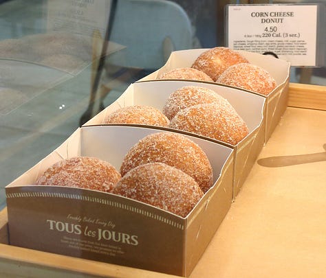 Tous les Jours display case items including fruit-loaded danishes, corn cheese donuts coated in granulated sugar, and a variety of croquettes including mango and chocolate