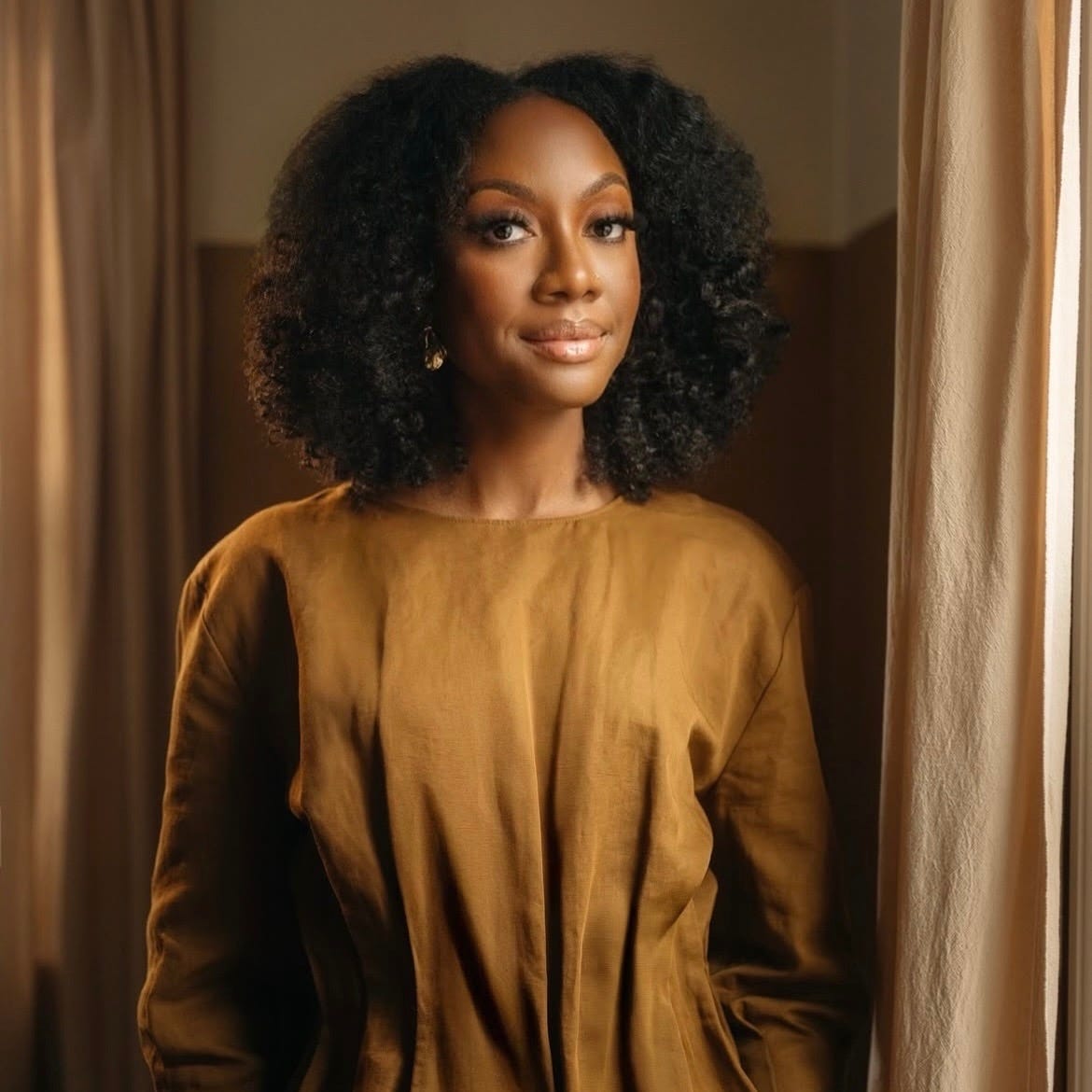 Professional portrait photograph of a Black woman with voluminous natural curly hair wearing gold hoop earrings and a mustard-gold colored long-sleeved top. She has a warm, confident smile and is photographed against a soft beige curtain backdrop with natural lighting creating a warm, professional atmosphere.