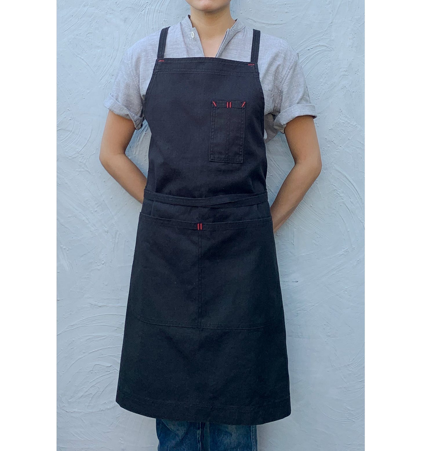 A person wearing a black apron, standing against a wall. A person wearing a black apron, standing against a wall.