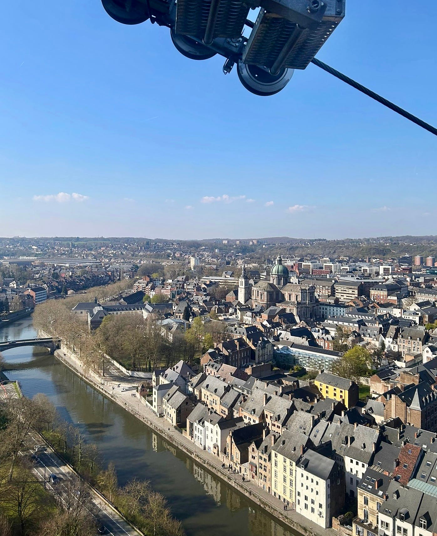 Aerial view of Namur, Belgium, taken from beneath a cable car gondola. The Meuse river runs through the centre of the frame, with the town's rooftops, a cathedral spire, and surrounding hills visible under a clear blue sky.