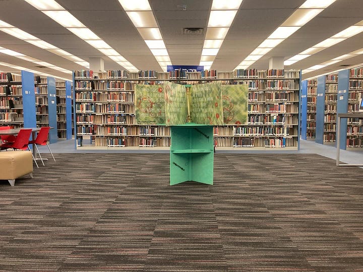 A big book with colorful pages sits on a display stand in front of a pair of windows. A painted, oversized book sits on a green display stand with library shelves visible behind it. A man stands next to an oversized book that is painted in red and green.
