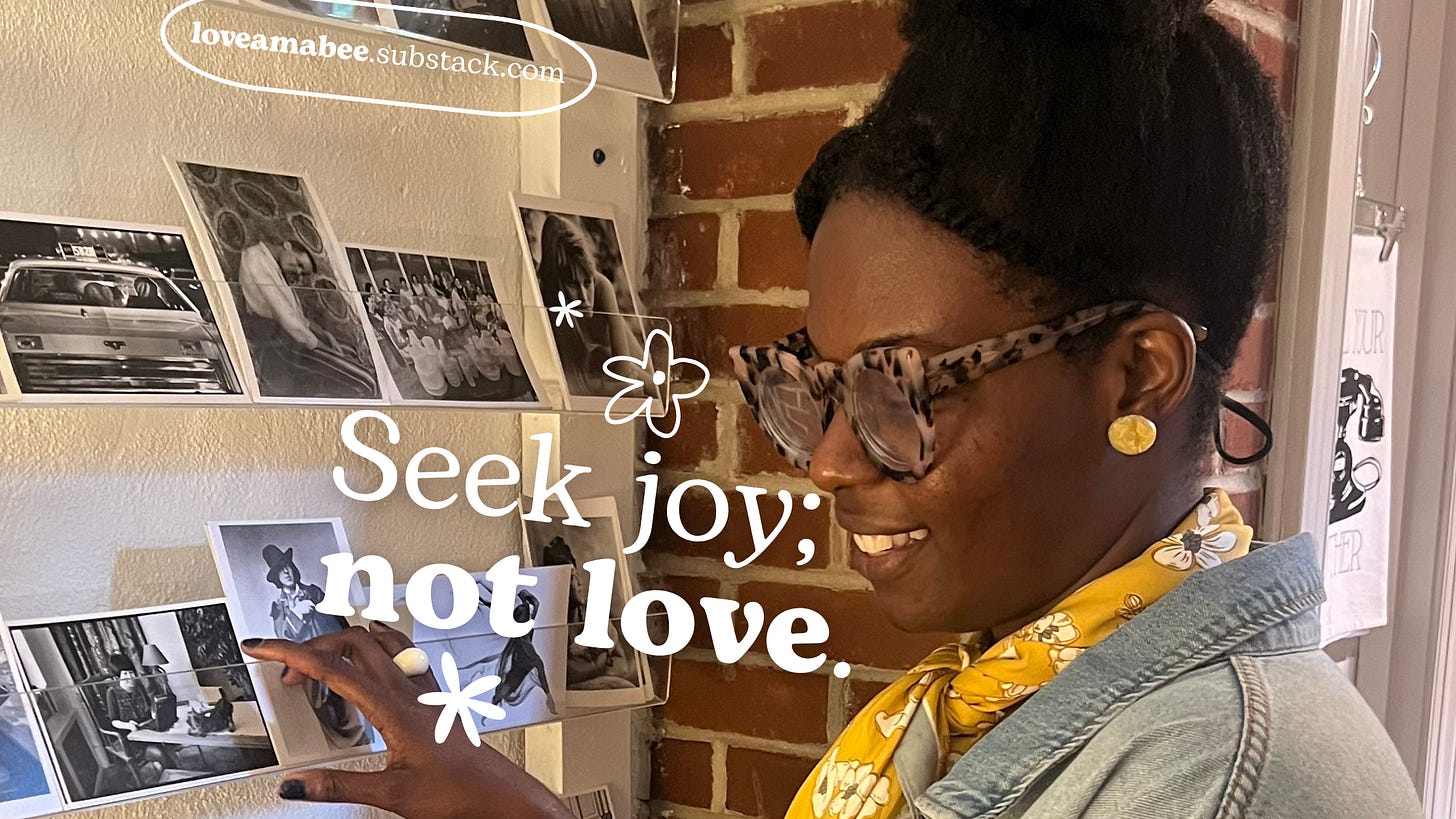 Seek joy, not love. Young black woman standing in front of a case of postcards with a smile; wearing a jean jacket and yellow floral scarf. 