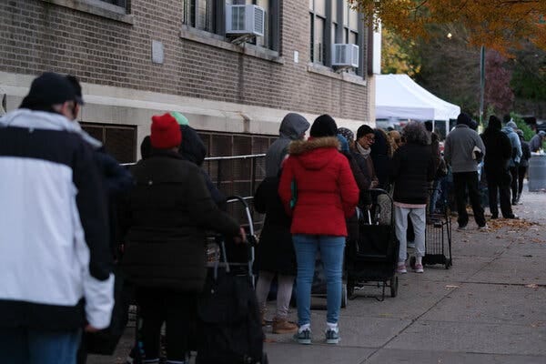 People, dressed in winter coats and towing grocery carts, wait in line at a food pantry.