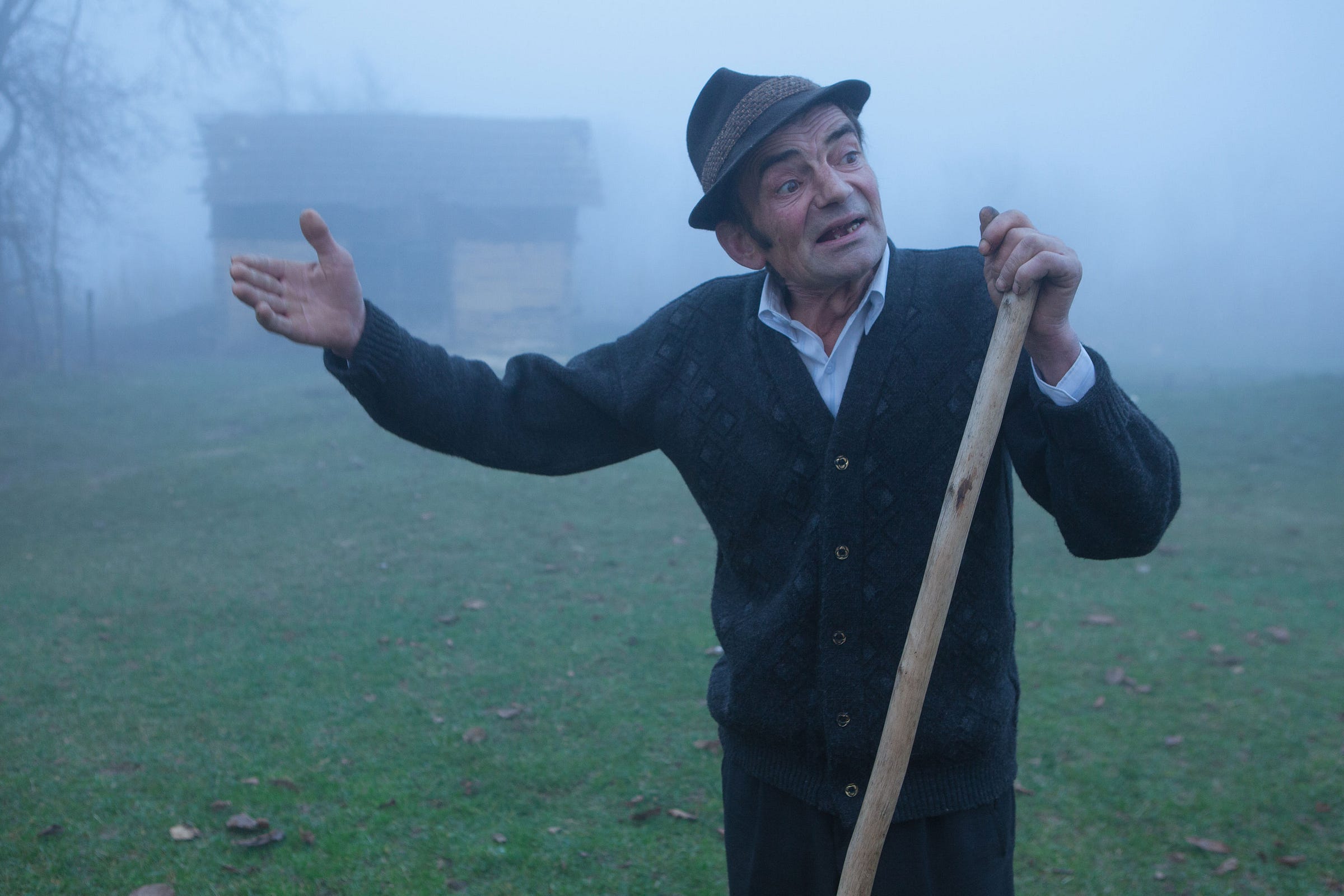 An older man wearing a hat and dark sweater stands on grass in a foggy field, holding a wooden stick and gesturing with his left hand. A rustic wooden building is faintly visible in the misty background.