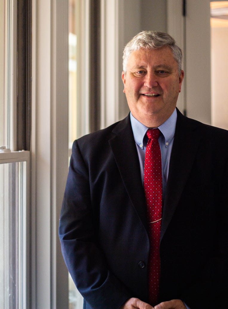 Austin Gardner Austin Gardner, in a suit and red tie, stands by a window, smiling warmly.