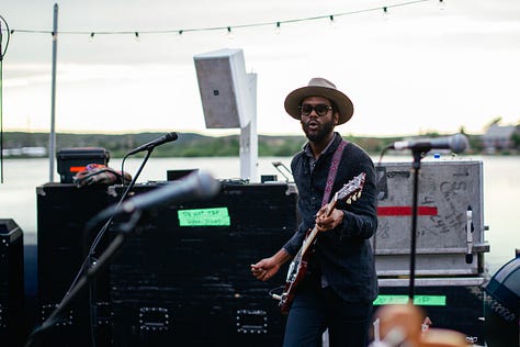 A gallery of three photos of Gary Clark Jr. and his band performing on an outdoor stage. The left photo shows him in silhouette against a bright orange sun, the middle photo shows him playing guitar, and the right photo shows him playing next to his bassist.
