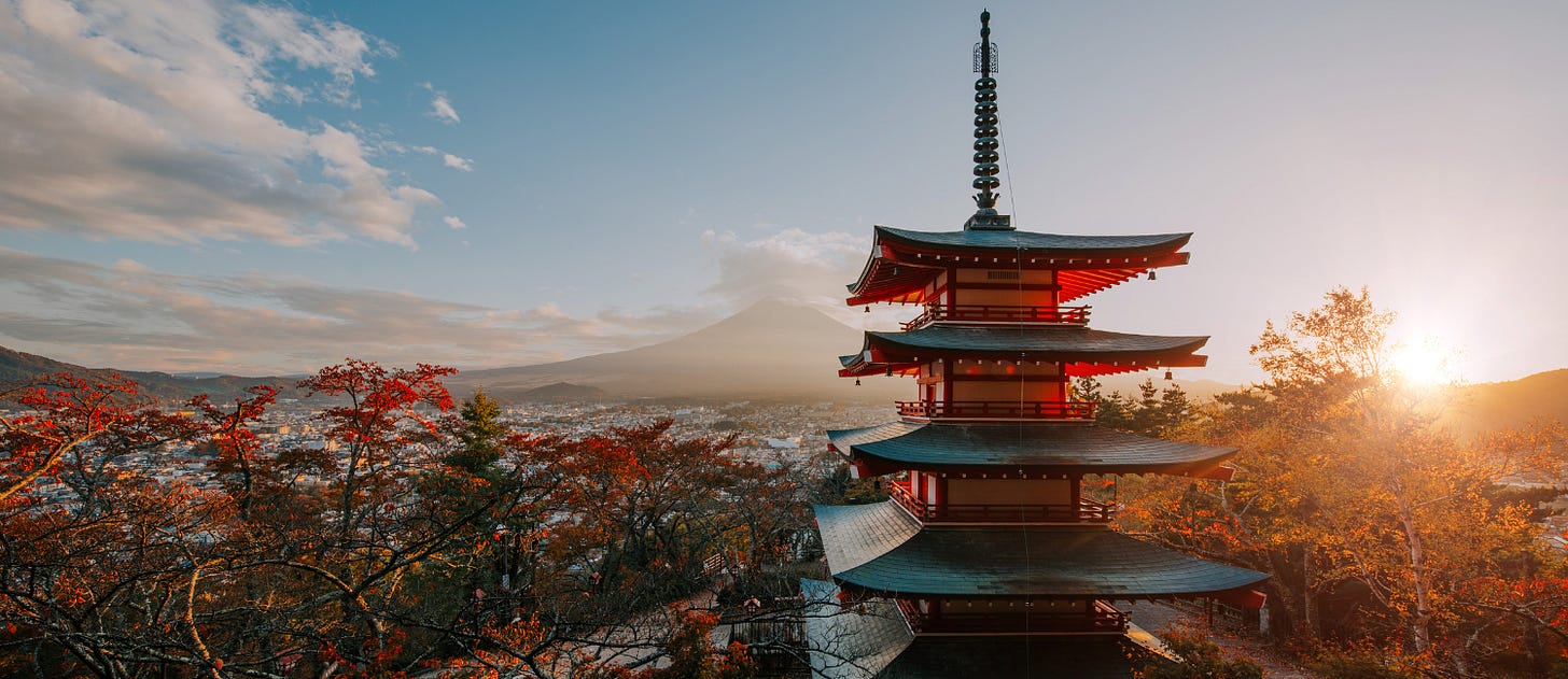 View of the five-story Chureito Pagoda in Fujiyoshida, Japan, surrounded by autumn foliage with red and orange leaves. In the background, Mount Fuji rises majestically under a soft haze, and the sun sets on the right side of the image, casting a warm golden glow across the sky and landscape.