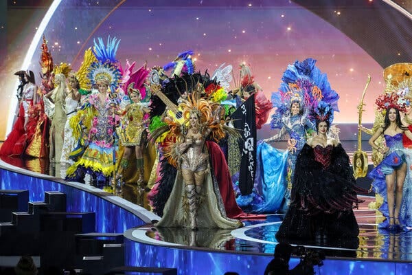 Women in colorful outfits, many with headdresses, walk on a stage.