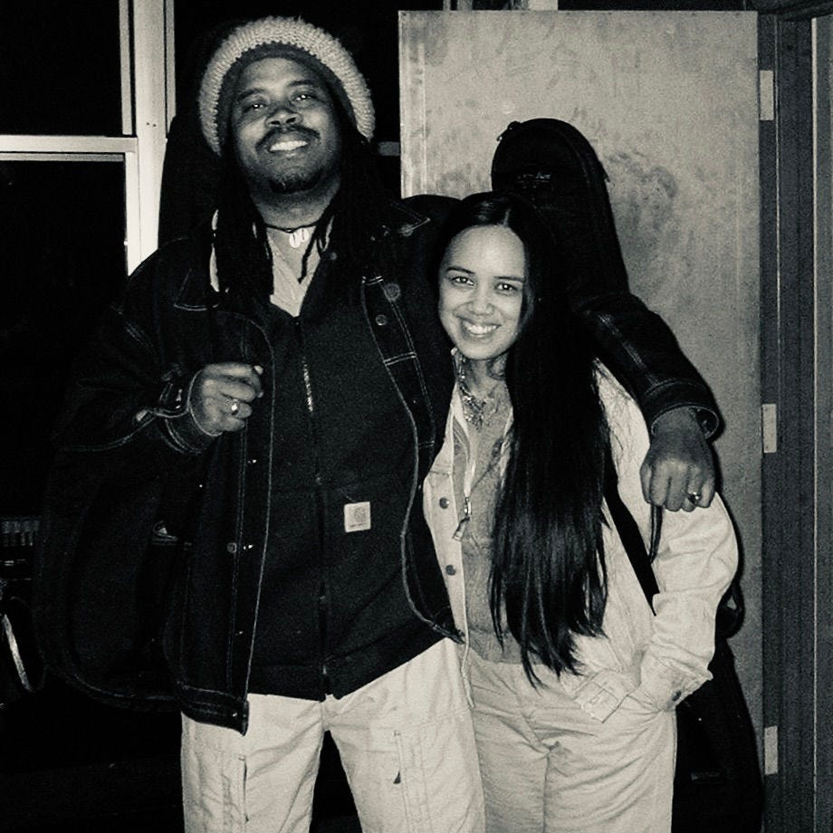 A man and woman smiling, arm in arm. The man wears a beanie; the woman has long hair. They're in a room with a window and a door. Black and white.