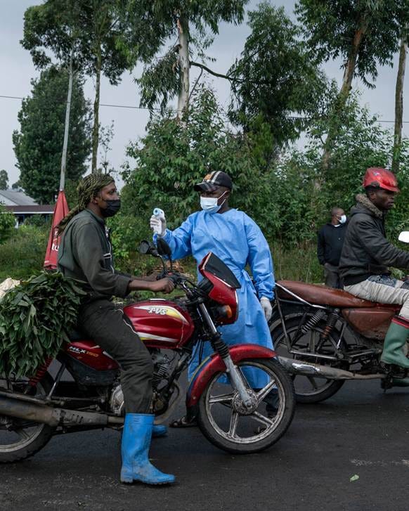A person in blue protective gear holds a thermometer to the face of a person on a red motorcycle. The rider wears a face mask and blue boots.