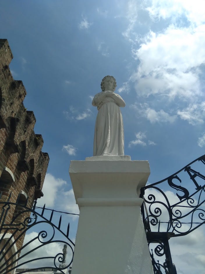 A statue of (presumably) an angel above a wrought iron cemetery gate. The scene is brightly lit but kind of unnerving.