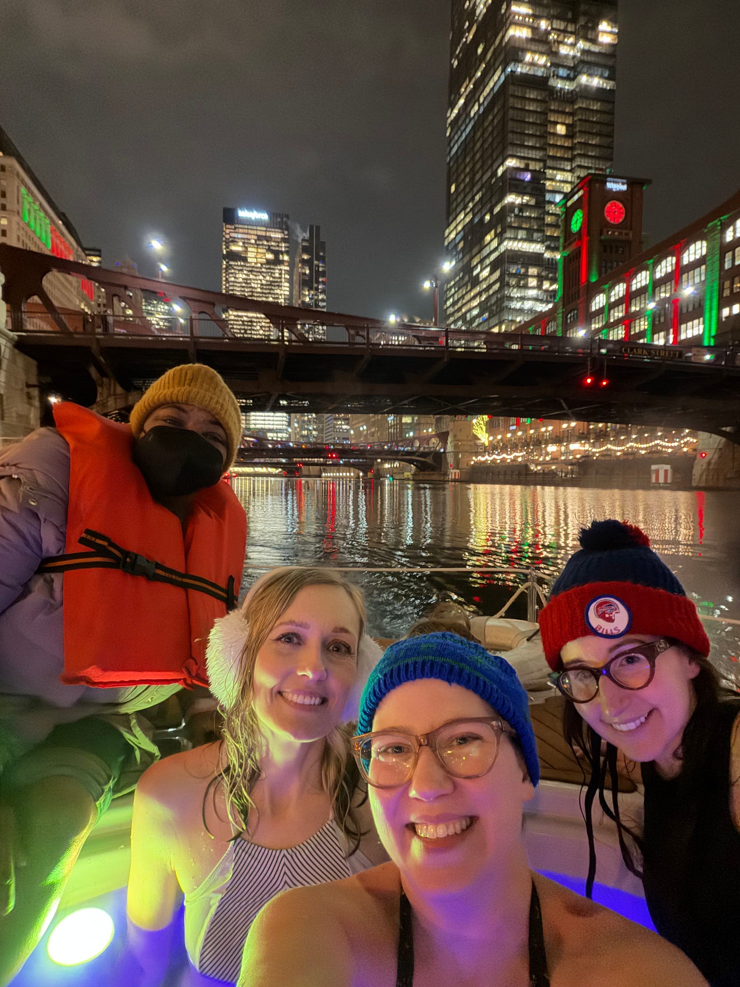 Four women in a hot tub boat in the Chicago River, buildings with red and green lights are seen in the background along with a couple of large bridges Four women in a hot tub boat in the Chicago River, buildings with red and green lights are seen in the background along with a couple of large bridges
