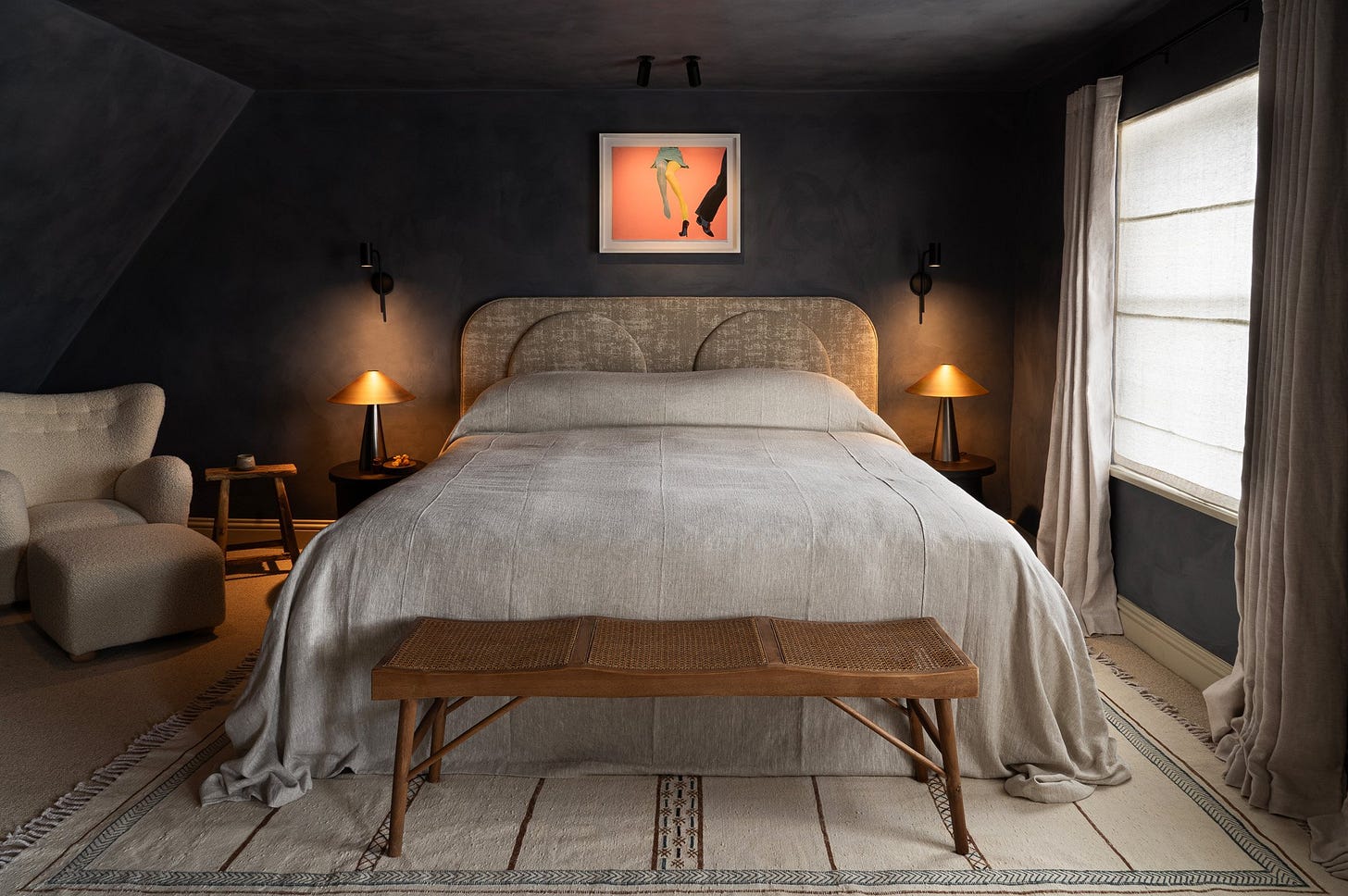 Bedroom with dark gray walls, beige bed with curved headboard, two wooden nightstands with matching lamps, framed artwork above the bed, a beige armchair with a matching ottoman, light beige curtains on a window, and a wooden bench at the foot of the