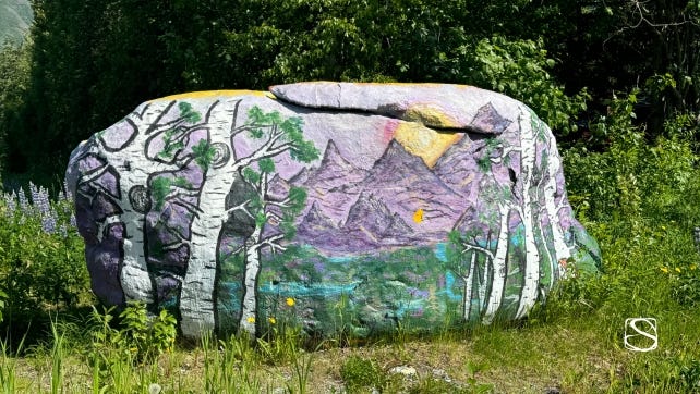Photo of large painted rock with nature landscape surrounded by plants and trees