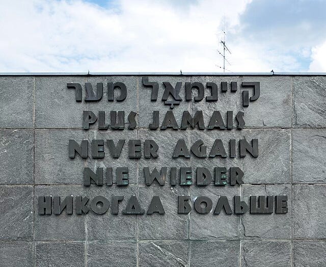 A stone wall reaches 80% up the image, a blue and white cloudy sky behind. On the slate grey wall in dark grey raised letters are the words "never again" in five languages - Hebrew, French, English, German, and Russian. A stone wall reaches 80% up the image, a blue and white cloudy sky behind. On the slate grey wall in dark grey raised letters are the words "never again" in five languages - Hebrew, French, English, German, and Russian.