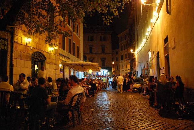 This may contain: people are sitting at tables in an alleyway with lights strung over the buildings on either side This may contain: people are sitting at tables in an alleyway with lights strung over the buildings on either side