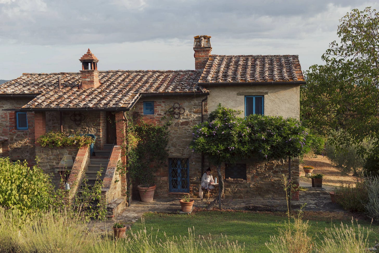 Charming rustic stone house with a tiled roof, lush greenery, and vibrant plants surrounding a staircase leading to a wooden door, set in the Tuscan countryside under a cloudy sky.