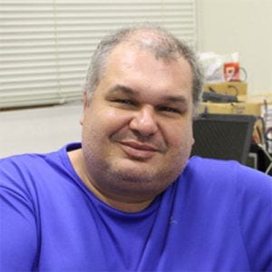 20260824_reynaldopinto homem de cabelos grisalhos usando camiseta azul e relógio no pulso, sentado em frente a mesa com laptop e equipamentos de medição, próxima a uma janela com persiana