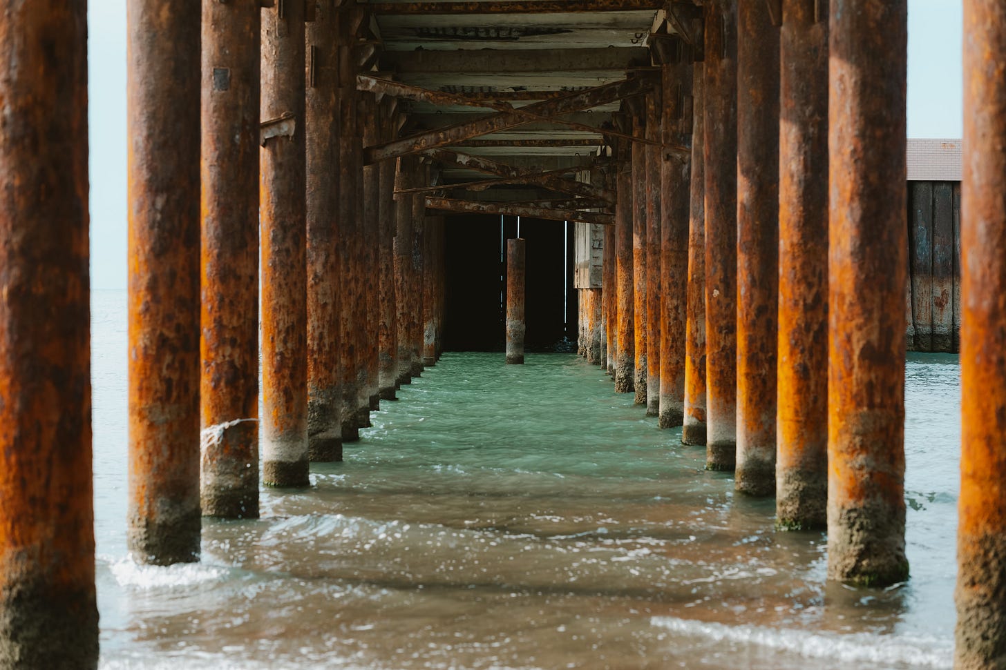 Representing the bridge we become between worlds and selves in a picture of an underneath view of a rustic pier in Baku, showcasing aged wooden beams over clear water