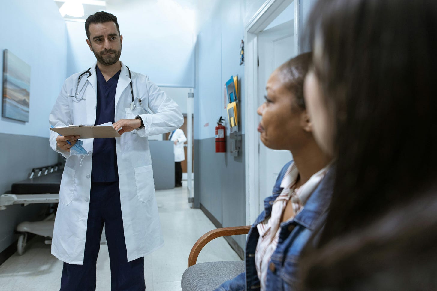 Stock photo: In a corridor in a hospital a young white male doctor in his twenties, wearing a lab coat, with a stethescope, stands holding a clipboard, looking at two women sitting on chairs; one woman is Black and in her thirties, the other has dark hair.