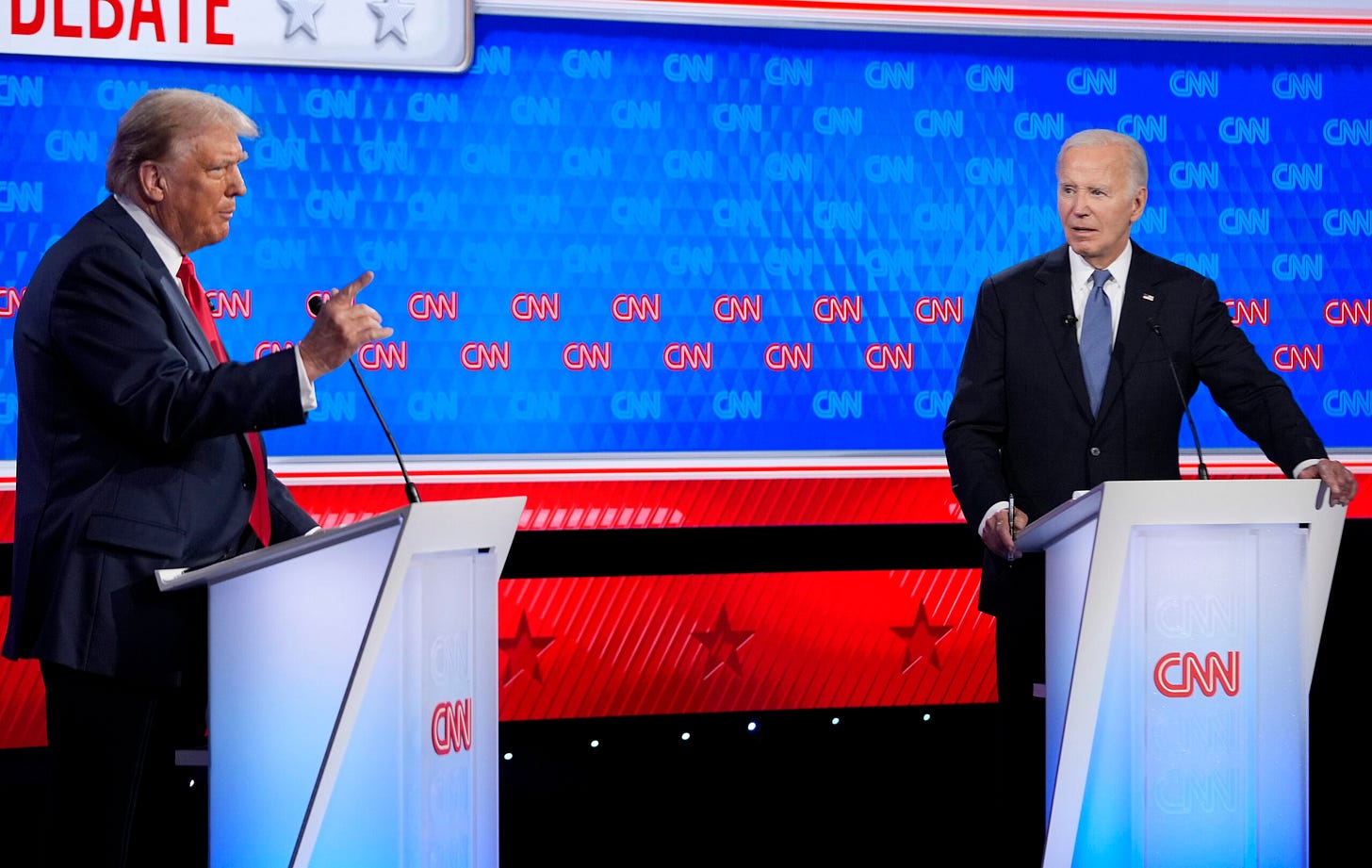 President Biden is standing on a podium on the right of the photo, former President Trump is on the left. Both are at CNN podiums with microphones. The background says CNN and is colored red, white, and blue with red stars at the bottom