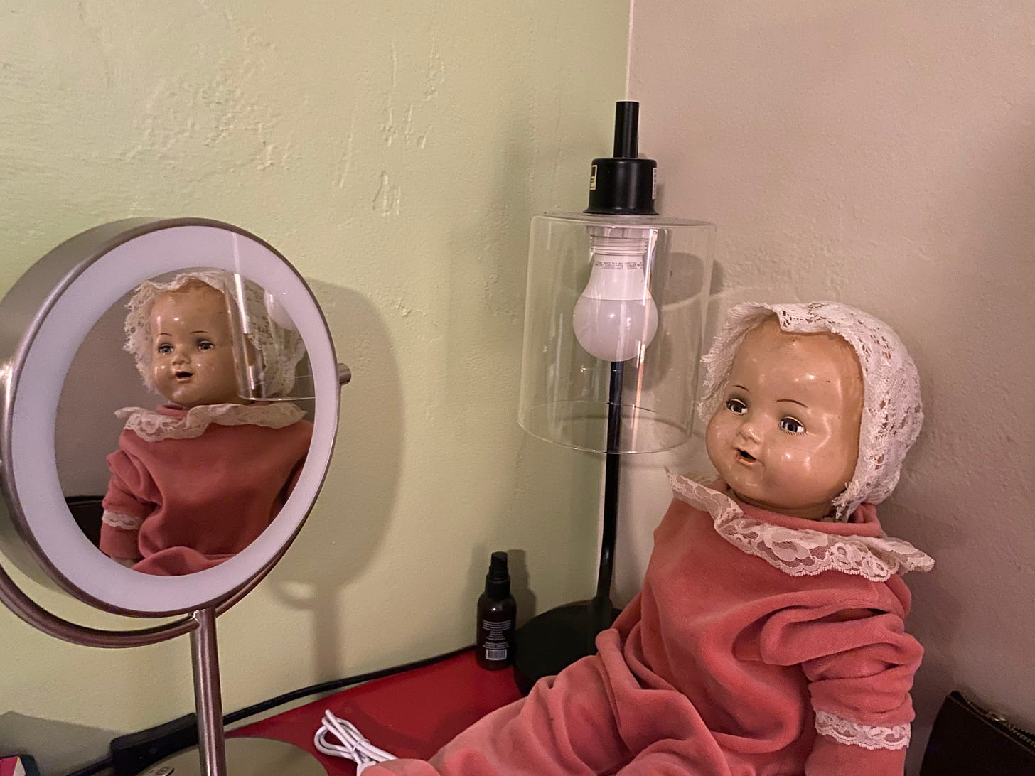 An old doll sits on a red desk, her reflection in a small makeup mirror. 
