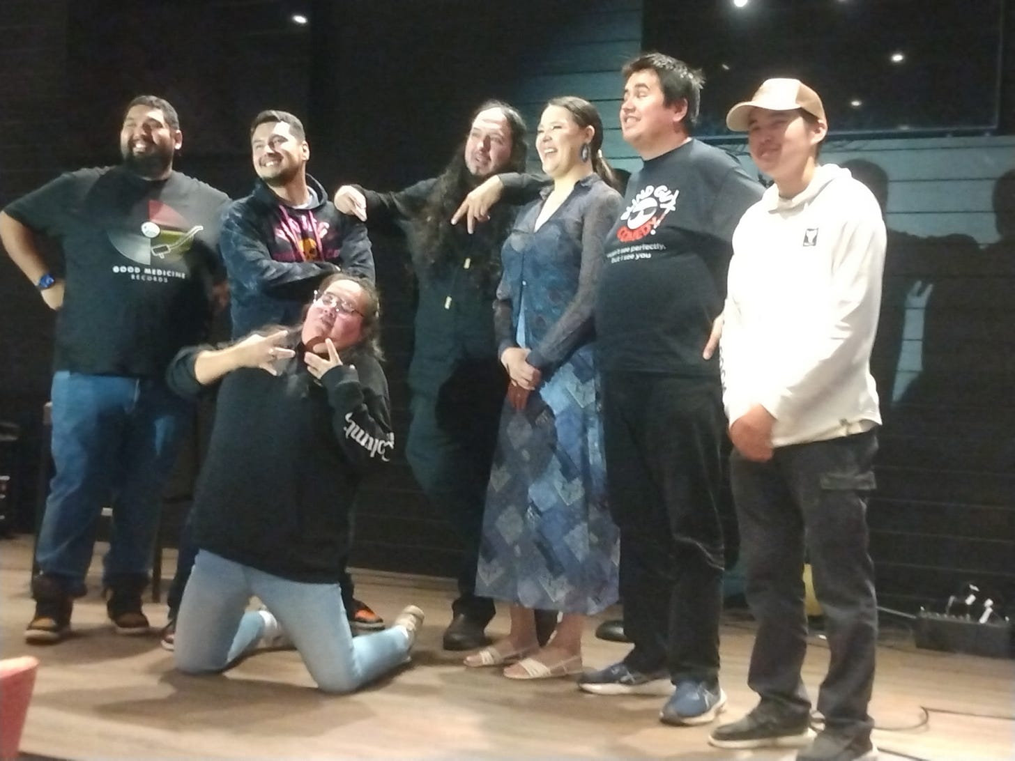 A group of seven people pose together on a small stage after a comedy show. Six people stand shoulder to shoulder, smiling toward the camera, while one person kneels playfully in front, making hand gestures. The group appears relaxed and happy, with casual clothing and warm expressions. The stage has a wooden floor, dark walls, and a large TV screen in the background.