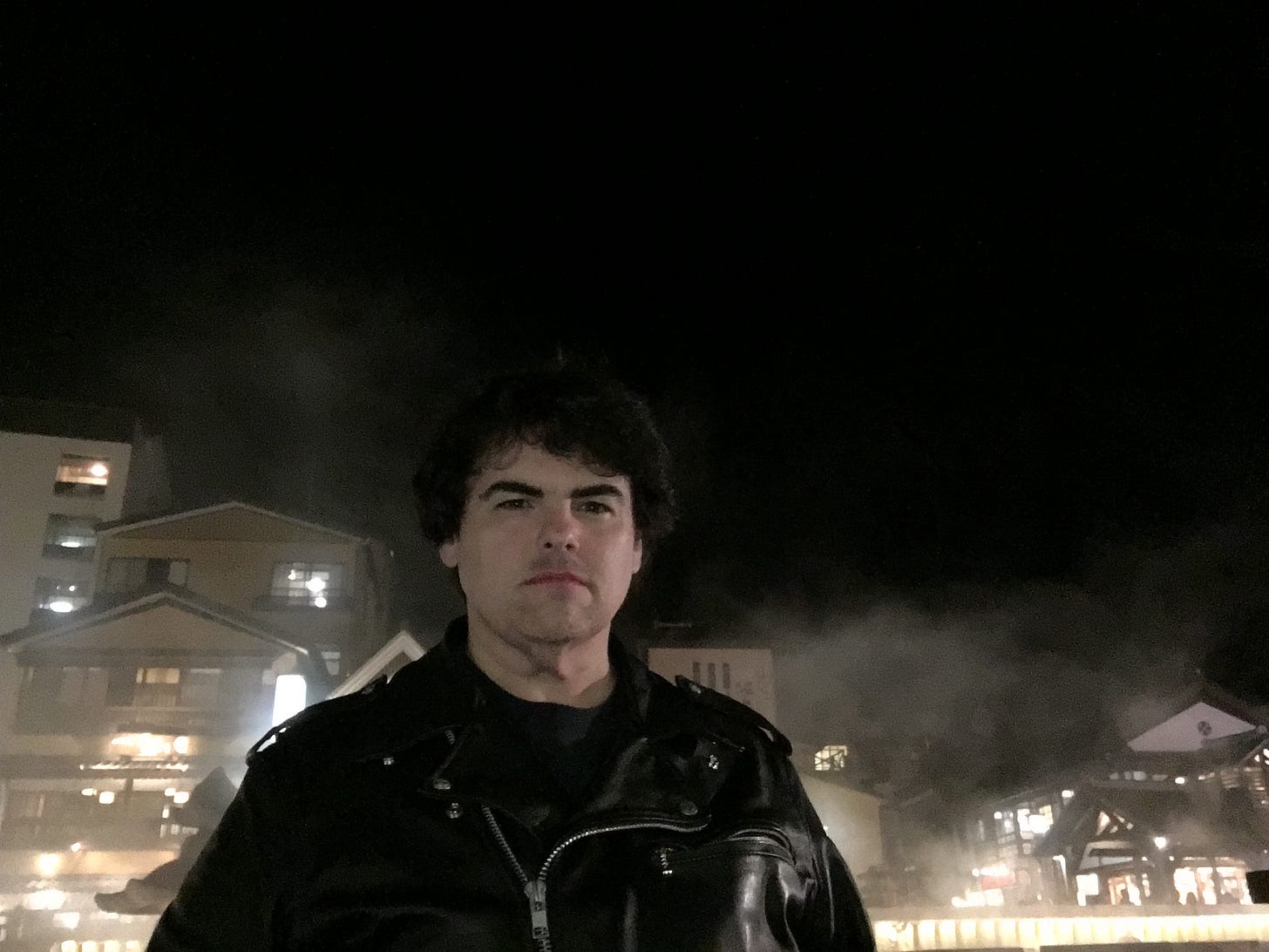 Male-appearing person with touseled black hair and a motorcycle jacket, surrounded by the steam of a Japanese onsen.  