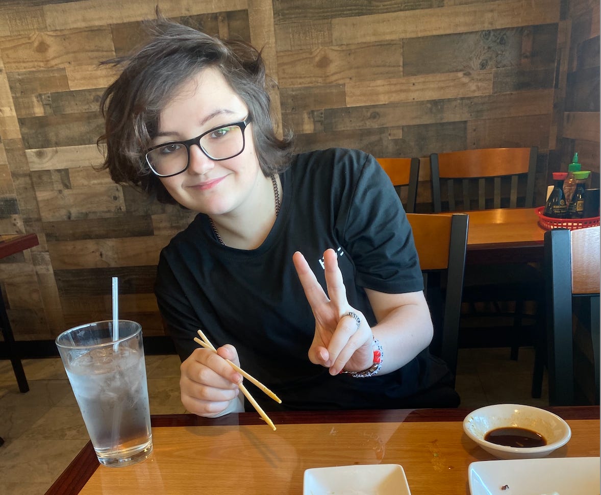 A teenager in glasses and a black t-shirt smiles at the camera, flashing a peace sign with one hand and holding chopsticks in the other.