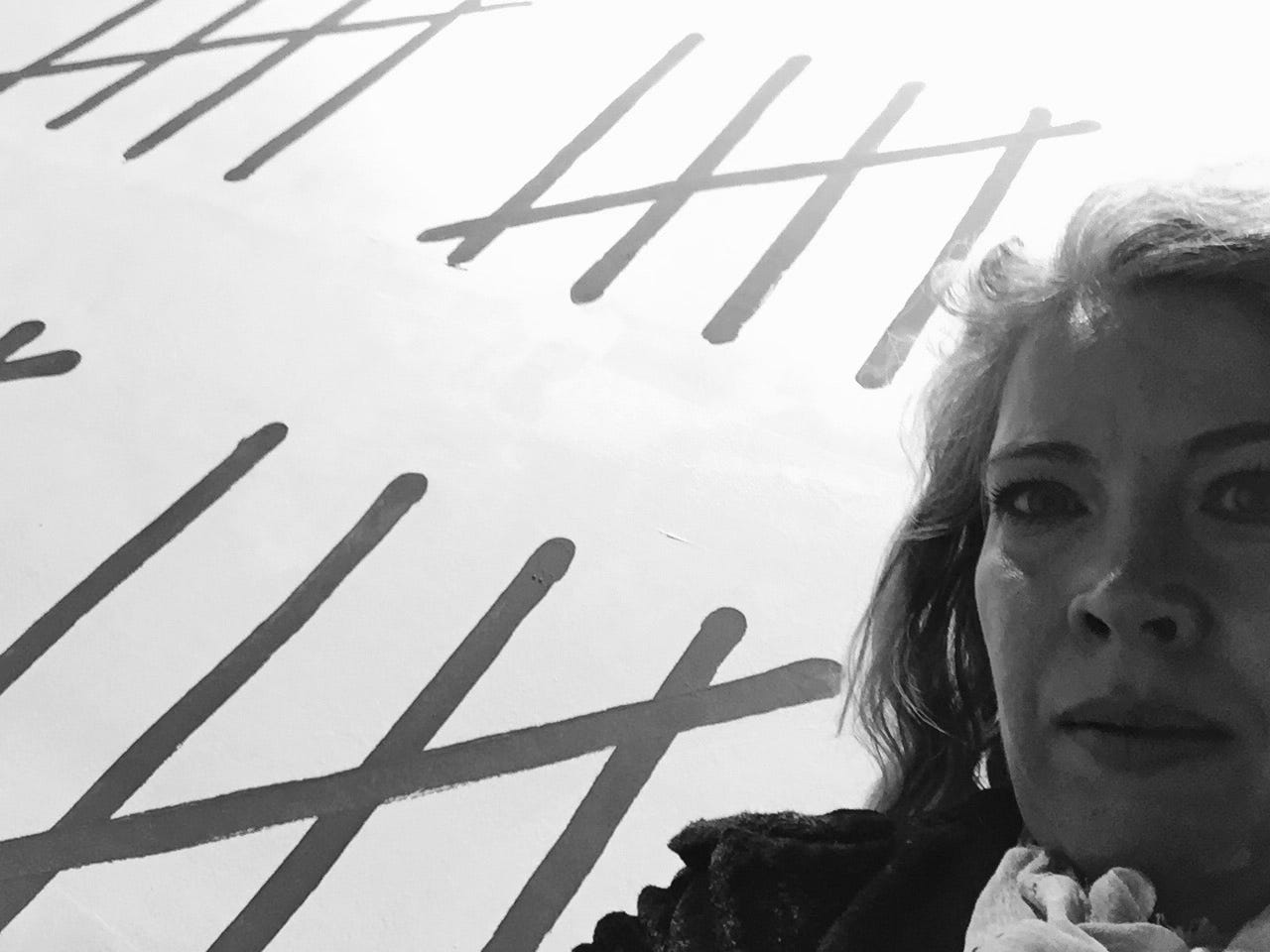 Black and white image of the author, Jennifer, standing in front of the Banksy mural. Three sets of hash marks are visible, only three quarters of Jennifers upper body and face are in the frame. She is wearing a dark fuzzy coat with a light scarf at her neck.