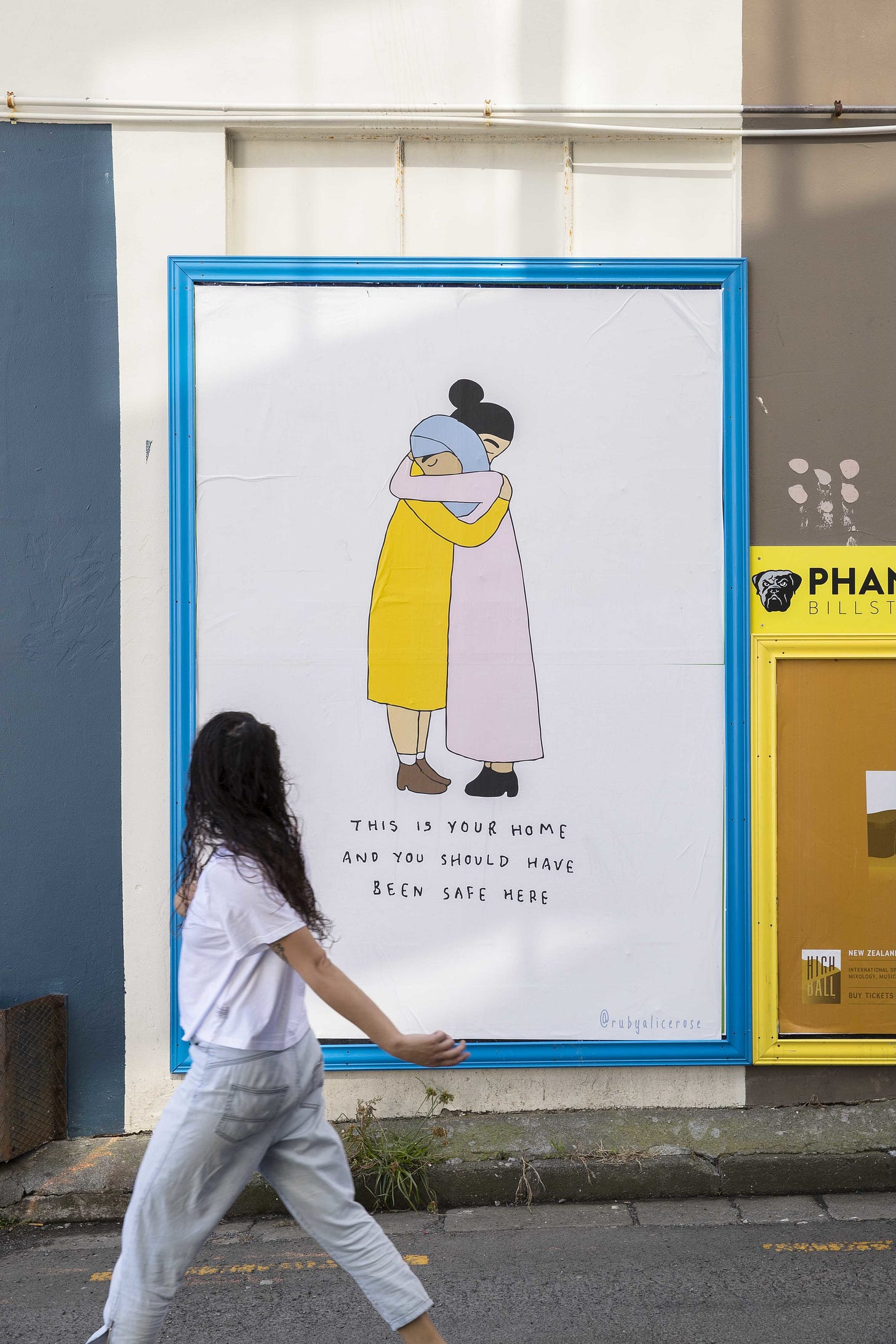 A poster put up in response to the Islamophobic terror attacks in Christchurch, Aotearoa New Zealand in 2019. In a blue frame a drawing of two women hugging, one in a lilac dress with pale blue hijab, the other in a yellow dress with dark hair pulled in a bun. The text below reads: This is your home and you should have been safe here.