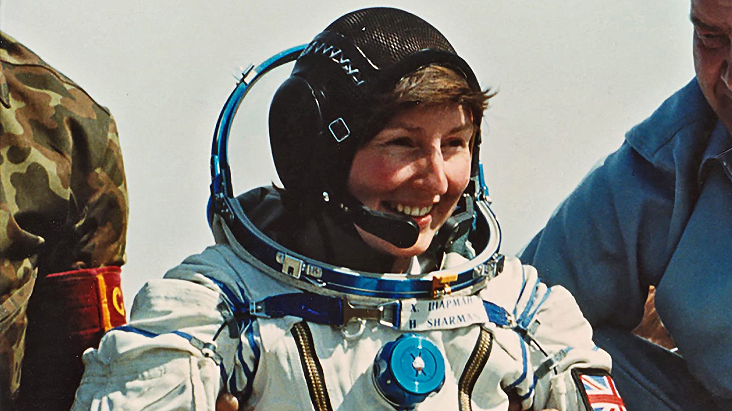 A woman wearing a space suit is helped out of a capsule