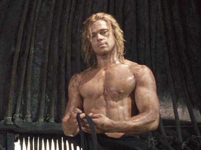 A shirtless man with long blond hair stands in a dimly lit room, holding fabric. His muscular build is highlighted, creating a serious mood.