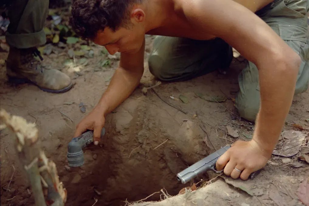 Public domain photo of American soldier, Sgt Ronald H. Payne, who was a Tunnel Rat during the Vietnam War, 1967, a year after author’s husband, Gregory, became one of the first Tunnel Rats. He has no shirt and is leaning over a square opening of a tunnel holding a .45 and a flashlight. Public domain photo of American soldier, Sgt Ronald H. Payne, who was a Tunnel Rat during the Vietnam War, 1967, a year after author’s husband, Gregory, became one of the first Tunnel Rats. He has no shirt and is leaning over a square opening of a tunnel holding a .45 and a flashlight.