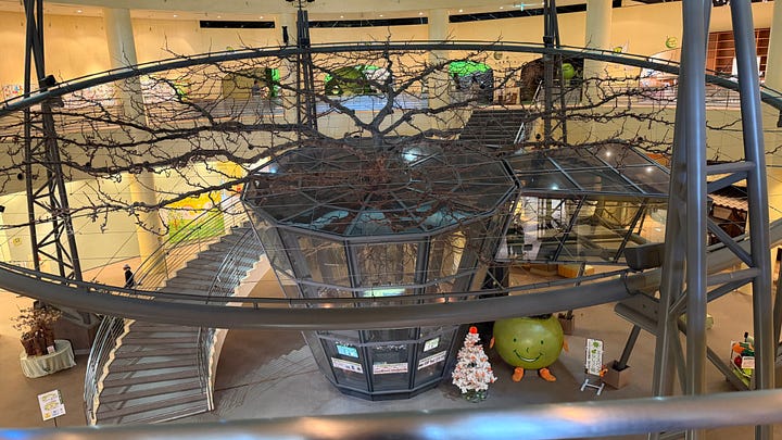 Image 1 (indoor scene): Alt text: Interior of a large museum atrium with a circular metal walkway surrounding a glass-walled central structure topped by branching tree-like sculptures. Curved staircases descend on the left, and colorful displays and mascots are visible on the lower floor.  Image 2 (outdoor scene): Alt text: View of a wide sandy dune stretching along a coastline with calm blue ocean behind it, seen from above green trees and buildings under a partly cloudy sky.