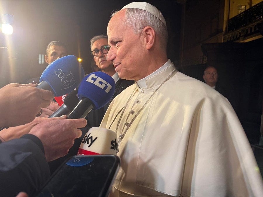 Elderly man with short white hair and glasses wears white zucchetto and white cassock with embroidered details stands in dimly lit outdoor area at night surrounded by journalists holding microphones labeled Sky, CGI, 200, and others while gesturing with hand and speaking. Several reporters including one with glasses and another in dark clothing listen attentively. Background includes balcony railing and soft lighting. Elderly man with short white hair and glasses wears white zucchetto and white cassock with embroidered details stands in dimly lit outdoor area at night surrounded by journalists holding microphones labeled Sky, CGI, 200, and others while gesturing with hand and speaking. Several reporters including one with glasses and another in dark clothing listen attentively. Background includes balcony railing and soft lighting.
