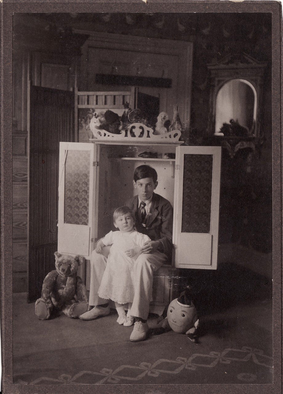 A vintage photograph from around 1930; it's black and white and depicts two children posing inside a cupboard. It's a formal portrait and there are teddy bears and toys arranged around them. A vintage photograph from around 1930; it's black and white and depicts two children posing inside a cupboard. It's a formal portrait and there are teddy bears and toys arranged around them.
