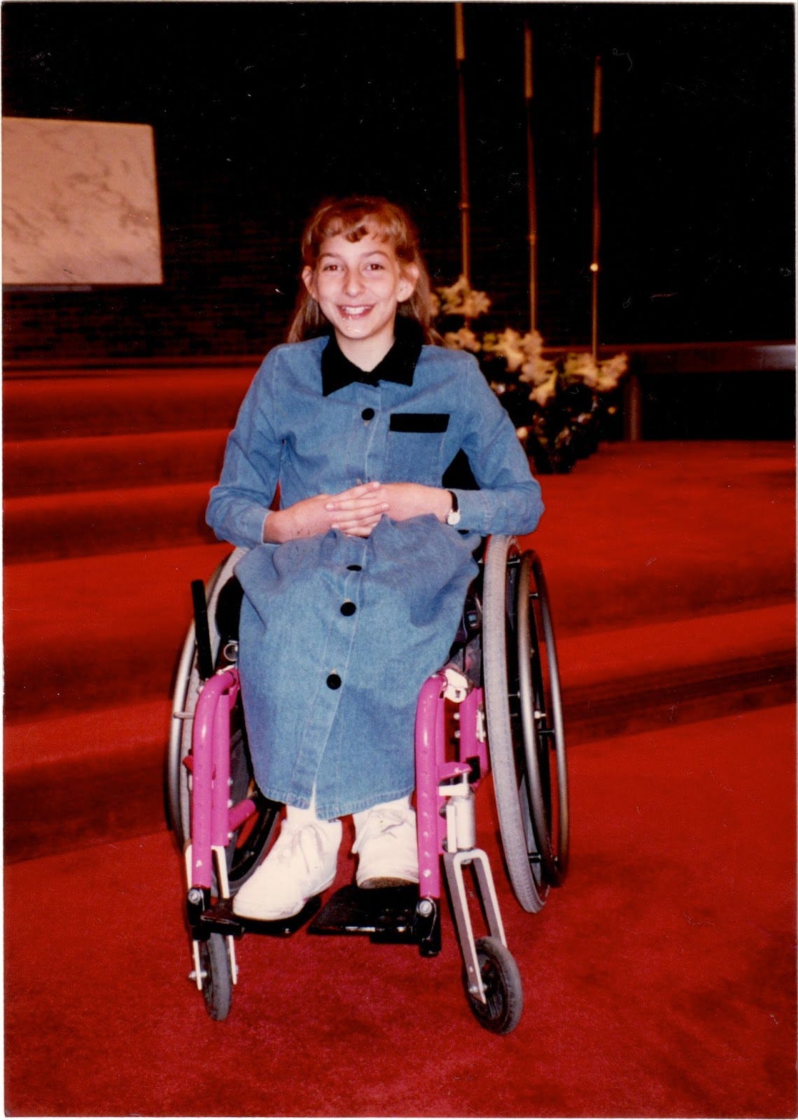 A much younger Rebekah, about 9 years old, sits in her hot pink wheelchair, her hands clasped awkwardly, anxiously in her lap. Her smile looks forced and uncomfortable. Her bangs are bouncy, her dress long and denim. Behind her are the red velvet steps leading up to the front of a church sanctuary. A much younger Rebekah, about 9 years old, sits in her hot pink wheelchair, her hands clasped awkwardly, anxiously in her lap. Her smile looks forced and uncomfortable. Her bangs are bouncy, her dress long and denim. Behind her are the red velvet steps leading up to the front of a church sanctuary.