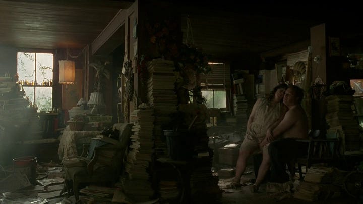 A woman sits on a man's lap inside a delapidated house filled with books, paper and clutter.