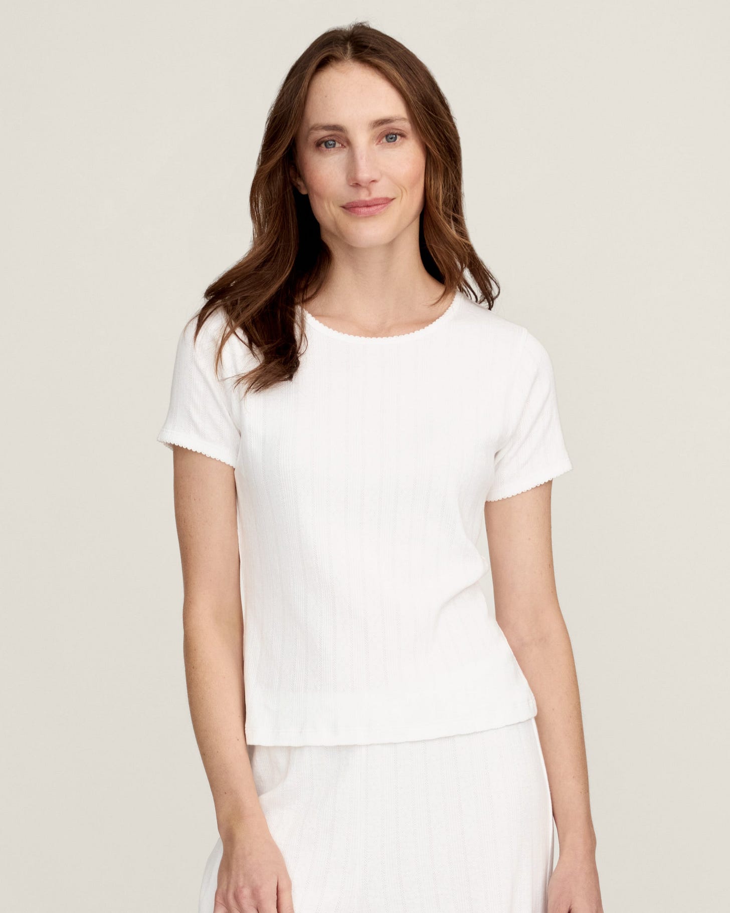 A woman with long brown hair wears the Women's Pointelle T-Shirt in White, standing against a plain light background and smiling gently at the camera.